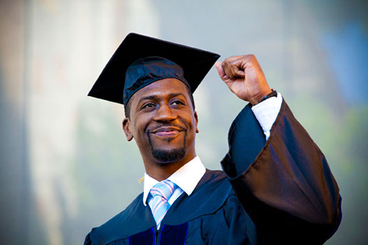 A male student in commencement regalia smiles and holds his fist in a victory gesture