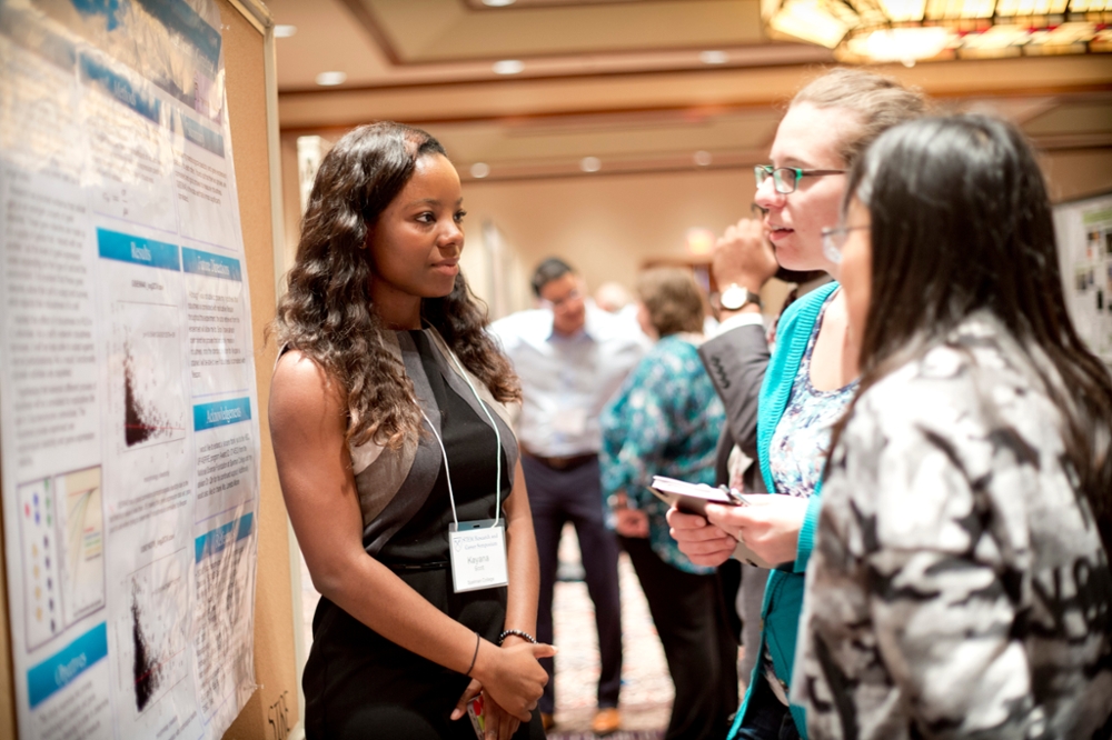 Three women conversing in front of a research poster