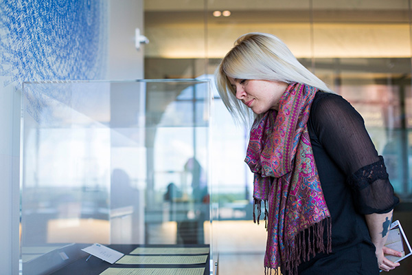 A woman with blonde hair and multicolored scarf around her next reads documents displayed in a glass museum display.